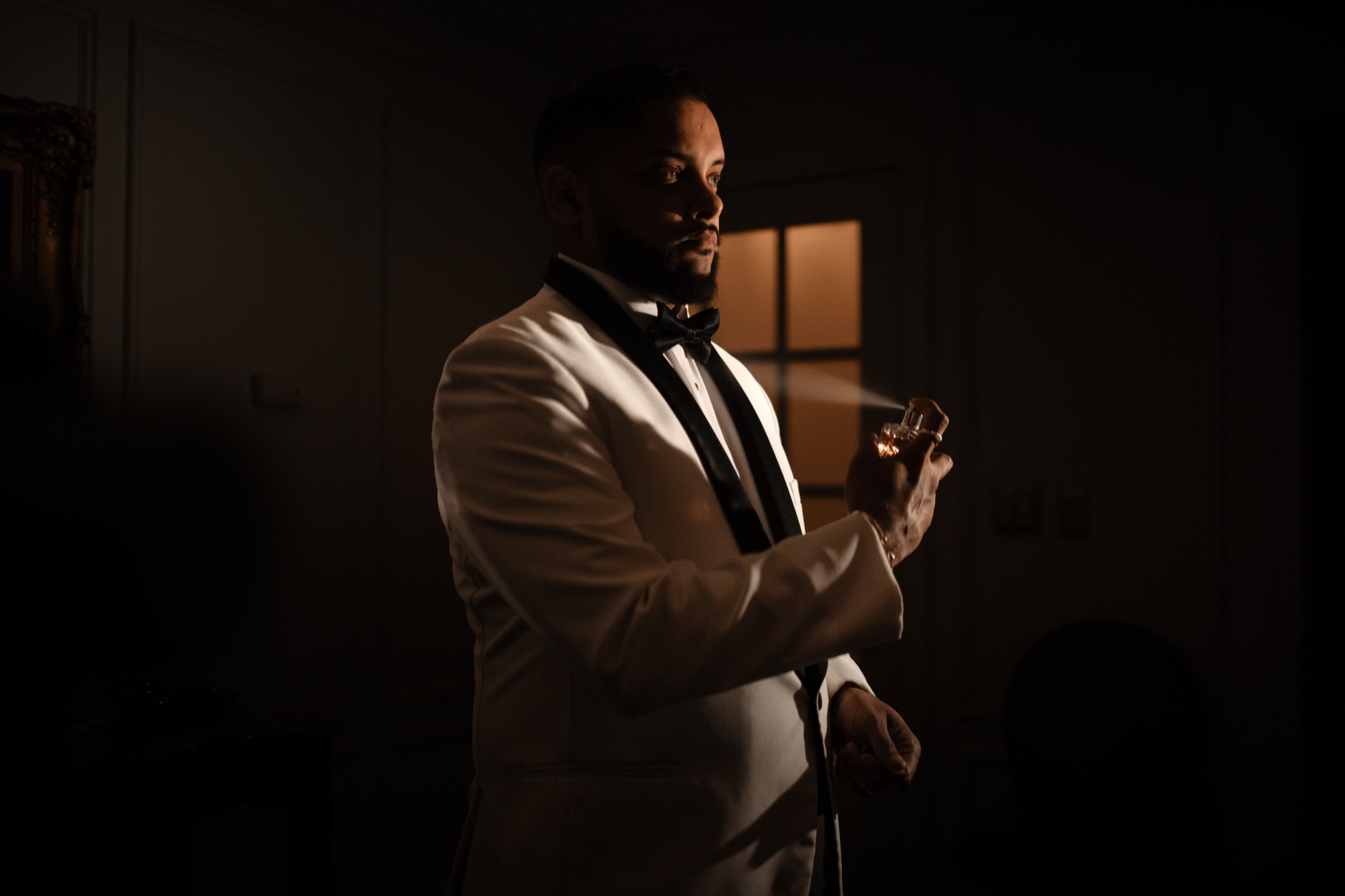 A man in a white dinner jacket and bow tie sprays fragrance in a softly lit room